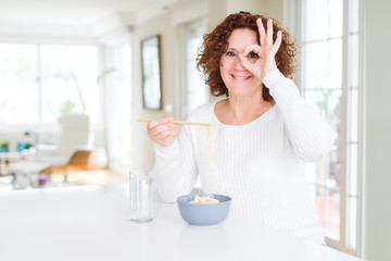Obraz na płótnie Canvas Senior woman eating asian noodles using chopsticks with happy face smiling doing ok sign with hand on eye looking through fingers