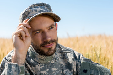 positive soldier in military uniform touching cap and smiling in field