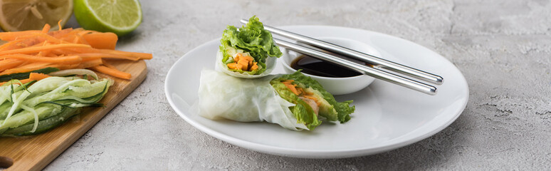 panoramic shot of tasty and served spring rolls with soy sauce on white plate with metal sticks