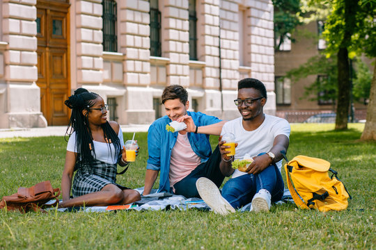 Group Of Happy Students Enjoying Picnic On Green Lawn Of School Yard, Drinking Fruit Drinks And Eat Cheeseburger . Multiethnic Friends Sharing Ideas During Free Time Outdoor.