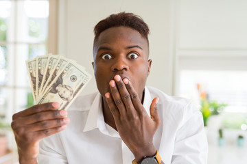 African american man holding twenty dollars bank notes cover mouth with hand shocked with shame for mistake, expression of fear, scared in silence, secret concept