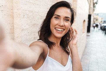 Portrait of happy woman smiling and taking selfie photo while standing over wall