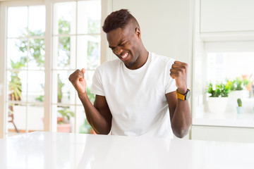 Handsome african american man on white table at home very happy and excited doing winner gesture with arms raised, smiling and screaming for success. Celebration concept.