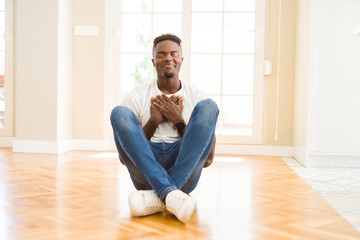 Handsome african american man sitting on the floor at home smiling with hands on chest with closed eyes and grateful gesture on face. Health concept.