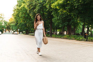 Portrait of cheerful woman smiling and holding cellphone while walking through green boulevard
