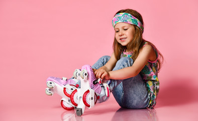 Little baby blond hair child sitting with roller skates in a white shirt and hairband happy smile on a pink studio background