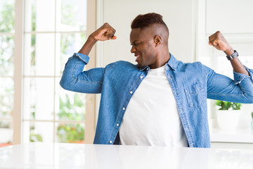 Fototapeta premium Handsome african american man at home showing arms muscles smiling proud. Fitness concept.