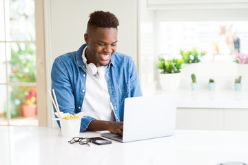 Handsome young african business man eating delivery asian food and working using computer, enjoying noodles smiling