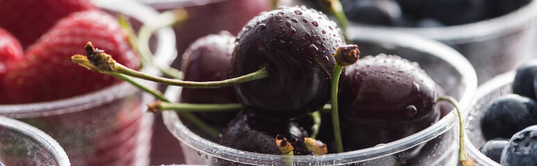 panoramic shot of fresh and ripe cherries with drops, blueberries and strawberries in plastic cups