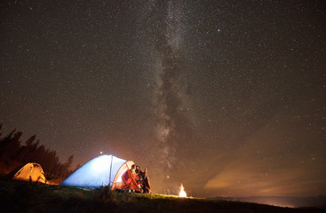 Night summer camping in the mountains. Young couple hikers having a rest together, sitting in the entrance tourist tent beside bonfire. Man pointing at night starry sky full of stars and Milky way.