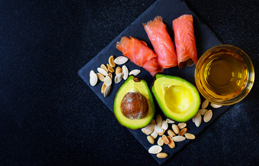 Selection of healthy fat sources, avocado, salmon, nuts, olive oil on a black plate. The concept of healthy eating. Top view, copy space, black background