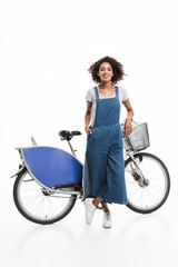Obraz premium Image of lovely african american woman dressed in denim overalls smiling while standing over bicycle