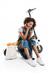 Image of cheerful african american woman using cellphone while sitting on electronic scooter with...