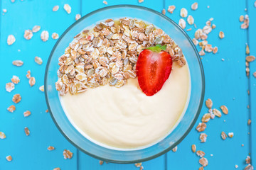 Healthy proper wholesome food. Yogurt with strawberries in a transparent deep plate on a blue wooden background, dry muesli scattered around