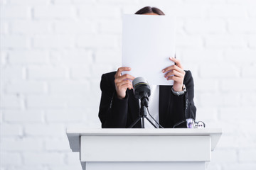 scared lecturer suffering from logophobia and hiding face behind paper while standing at podium...