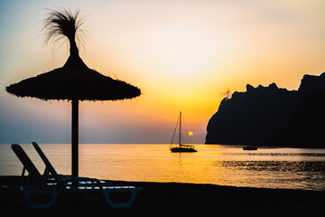 Mediterranean Sunrise off the Mountainous Coast of Mallorca in the Ballearic Islands with Silhouetted Sailboat and Beach Umbrella