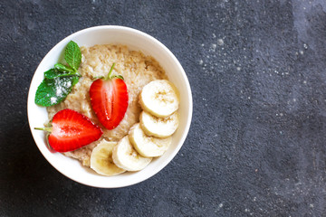 Fresh oatmeal with strawberries and banana slices in a white plate stands on a dark concrete background. On the right there is free space for inscriptions.