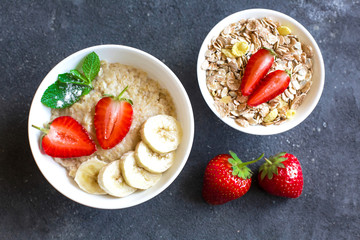 Oatmeal with strawberries and banana slices in a white plate standing on a dark concrete background. Decoration of dry oatmeal scattered around on the background and  strawberries.