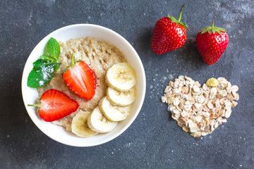 Oatmeal with strawberries and banana slices in a white plate standing on a dark concrete background. Decoration of dry oatmeal in the form of a heart on the background and strawberry slices.