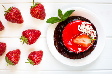 Red round cake with strawberries and mint leaves on a white saucer, which stands on a white wooden background to the right. On the left in bulk are the berries of fresh strawberries.