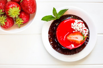 Red round cake with strawberries souffle decorated with mint leaves on a plate, which stands on a white wooden background. Nearby is a transparent bowl with fresh strawberries.