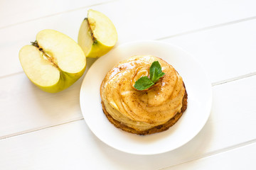 On a white plate lies a cake of apple charlotte with mint leaves close-up on a white wooden background. To the left of the plate lies an apple cut into pieces.