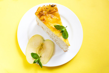 Cottage cheese pie with an apple  apples on a white plate stands on a yellow background. Nearby is a piece of apple decorated with mint leaves.