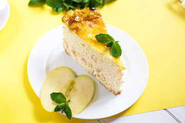 Cottage cheese pie with an apple from yellow and green apples on a white plate stands on a yellow background. Nearby is a piece of apple decorated with mint leaves.