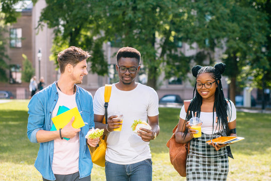 Group of three positive multiethnic students people coworkers talking, sharing ideas during lunch break standing outdoor university campus with books and backpacks. University education concept.