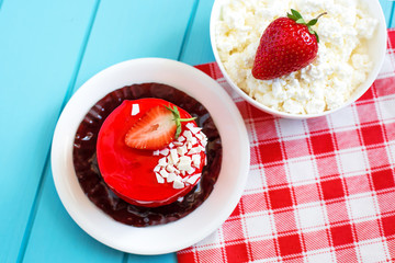 Red round cake with strawberry souffle, decorated with mint leaves on a plate, which stands on a blue wooden background. Nearby is a plate with cottage cheese and strawberries.