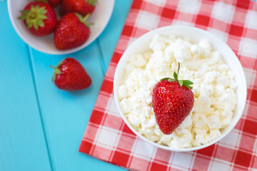 Fresh tasty granular cottage cheese with strawberries in a white deep plate on a blue wooden background and a cotton towel in a red cell, strawberries 
