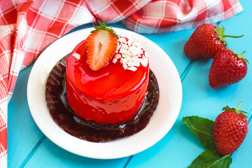 Red round cake with strawberry souffle, decorated with mint leaves on a plate, which stands on a blue wooden background. Nearby are strawberries.