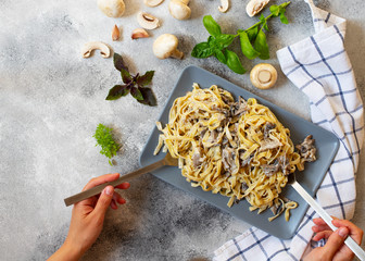 Homemade Italian fettuccine pasta with mushrooms and cream sauce served on a gray plate (Fettuccine al Funghi Porcini). Female's hands serving pasta. Gray background, top view, copy space