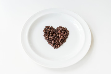Roasted coffee beans in a shape of heart in ceramic plate on a white background, top view