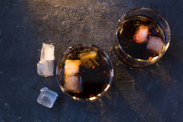 Two glasses with cocktails: rum cola and whiskey cola stand on a gray blue concrete textured background and there are a few ice cubes lying around