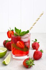 On a light gray background there is a strawberry cocktail in a glass with ice and mint, strawberries and slices of sliced lime