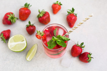 On a light background there is a strawberry cocktail in a glass with ice and mint, a bowl with strawberries and lime slices