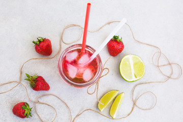 on a light gray ceramic background there is a glass with a fresh strawberry cocktail with ice and mint, next is a strawberry, lime slices and a rope around them