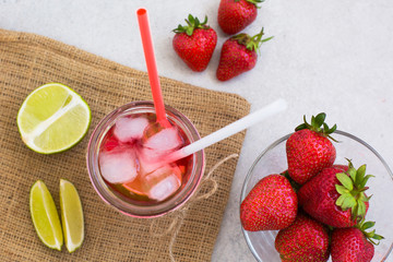 on sackcloth there is a glass with a fresh strawberry cocktail with ice and mint, next is a strawberry and lime slices