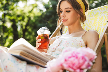 Concentrated beautiful young pretty woman in green park drinking juice lies on sun bed reading book.