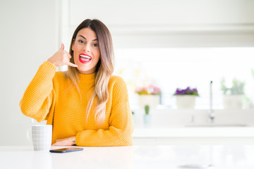 Young beautiful woman drinking a cup of coffee at home smiling doing phone gesture with hand and fingers like talking on the telephone. Communicating concepts.