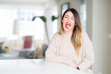 Young beautiful woman wearing winter sweater at home sticking tongue out happy with funny...