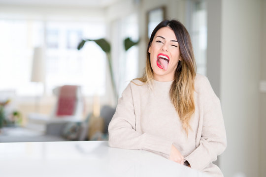 Young beautiful woman wearing winter sweater at home sticking tongue out happy with funny expression. Emotion concept.