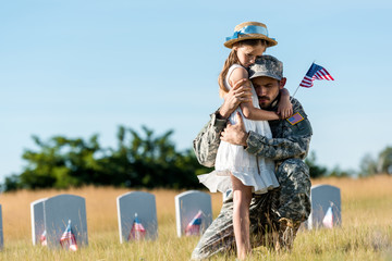 military man with closed eyes hugging daughter near headstones in graveyard - obrazy, fototapety, plakaty