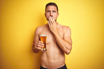 Fototapeta premium Young handsome shirtless man drinking a pint of beer over isolated yellow background cover mouth with hand shocked with shame for mistake, expression of fear, scared in silence, secret concept