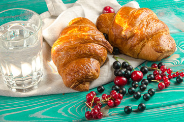 Fresh ruddy croissants with berries lie on a wooden table next to fresh black currant berries, red currants, cherries