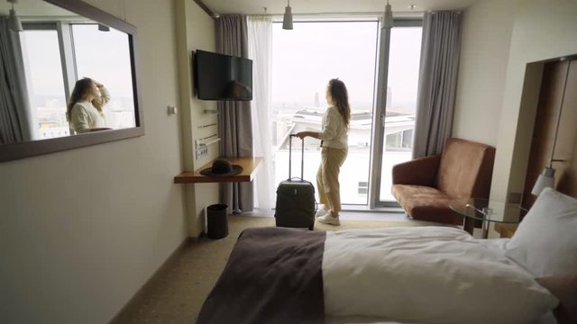  Women just arrived to hotel room. View from the back charming girl opening curtains in apartment. wide angle shot female with stretched out arm and suitcase enjoying view through big window.