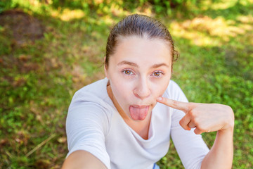 Young girl take selfie from hands with phone showing tongue and funny face sitting on park or garden background. Portrait of young attractive woman making selfie photo on smartphone in summer day