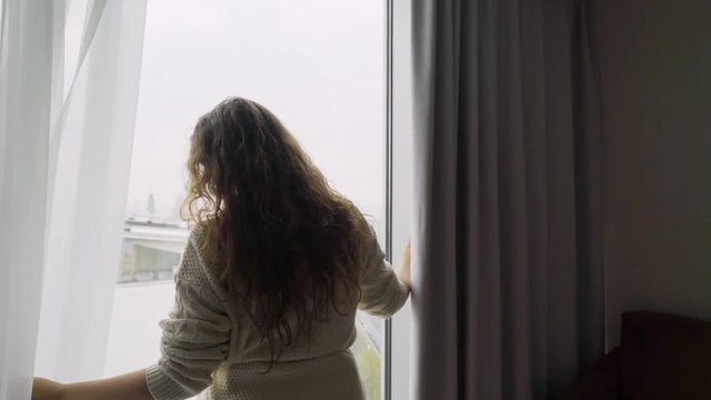 Young girl opening curtains in dark, apartment is illuminating bright light. Back view women just arrived hotel room, stretched out arm and enjoying view through big window.