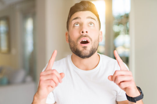 Young handsome man wearing casual white t-shirt at home amazed and surprised looking up and pointing with fingers and raised arms.